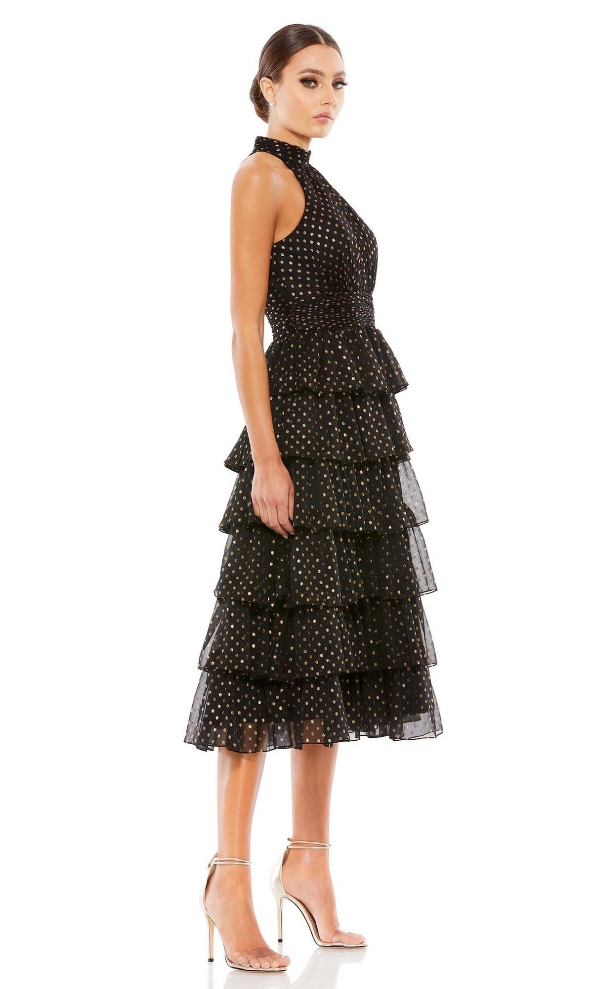 A woman in profile wears the Mac Duggal 49510 Midi Black Wedding-Guest Dress, a sleeveless tea-length gown with gold polka dots and tiered silhouette, paired with high heels against a white background.